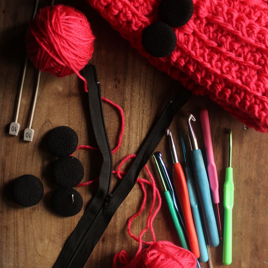 Red yarn, hooks, buttons and a zip laid out beside a half-finished piece